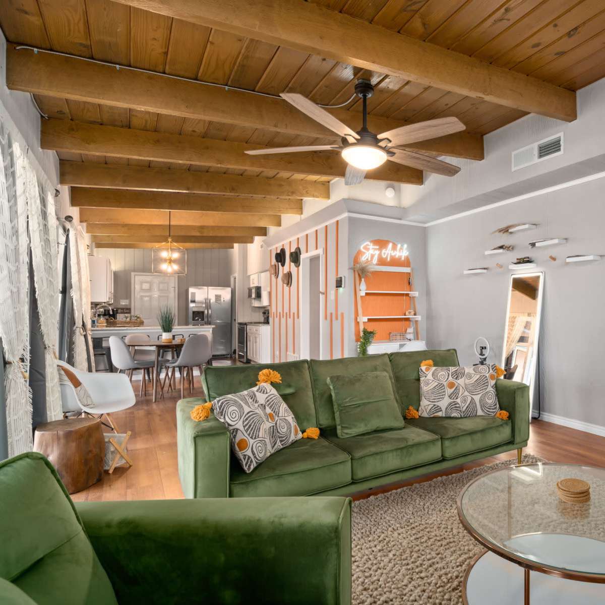 A living room with wooden beams on the ceiling, a green sofa adorned with decorative pillows, a glass coffee table, and an open layout leading to a kitchen.