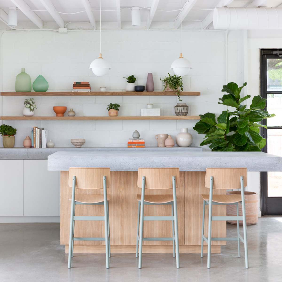 A modern kitchen features a central island with wooden base and concrete countertop, surrounded by minimalist chairs, with open shelving displaying decorative items and plants.