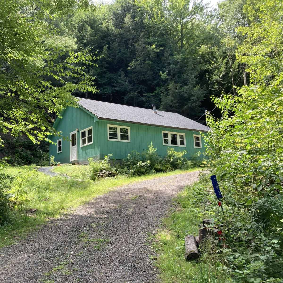 A green house is situated at the end of a gravel driveway, surrounded by lush greenery and trees.