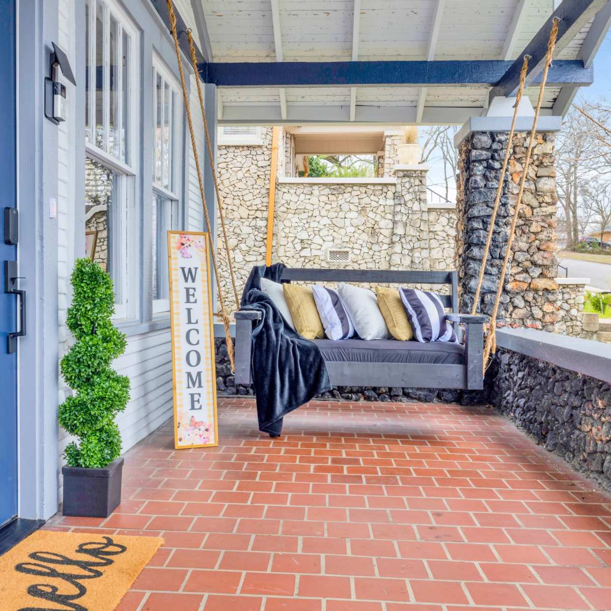 A porch with a blue front door, a welcome sign, a sofa adorned with pillows, a throw blanket, and potted plants.