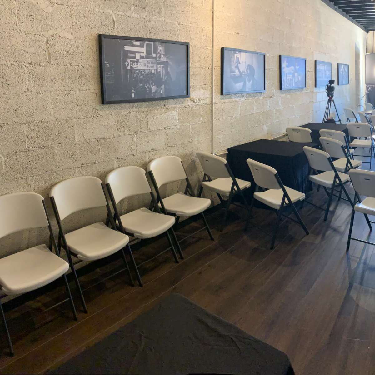An empty room set up with black tablecloths covering several tables and white chairs arranged in a meeting space, with framed photographs on the walls.