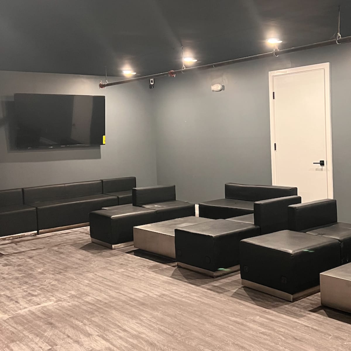 A minimalist lounge area features several black sofas and low tables arranged around a wall-mounted television and a white door.