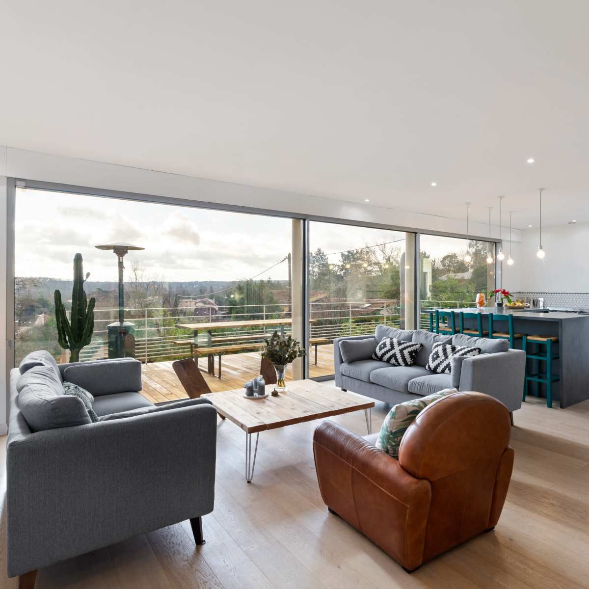 A modern living room with two gray sofas, an armchair, a coffee table, and a kitchen visible in the background, featuring large windows that overlook a scenic view.