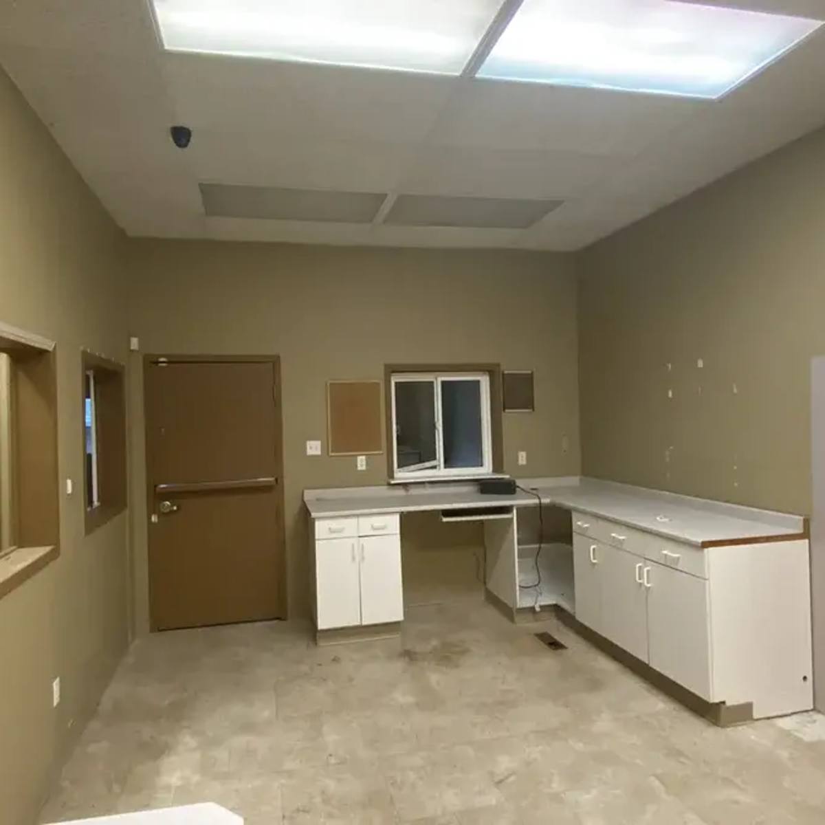 An empty, beige-walled room with a sink, countertops, and two windows, one of which is mounted in a wall.