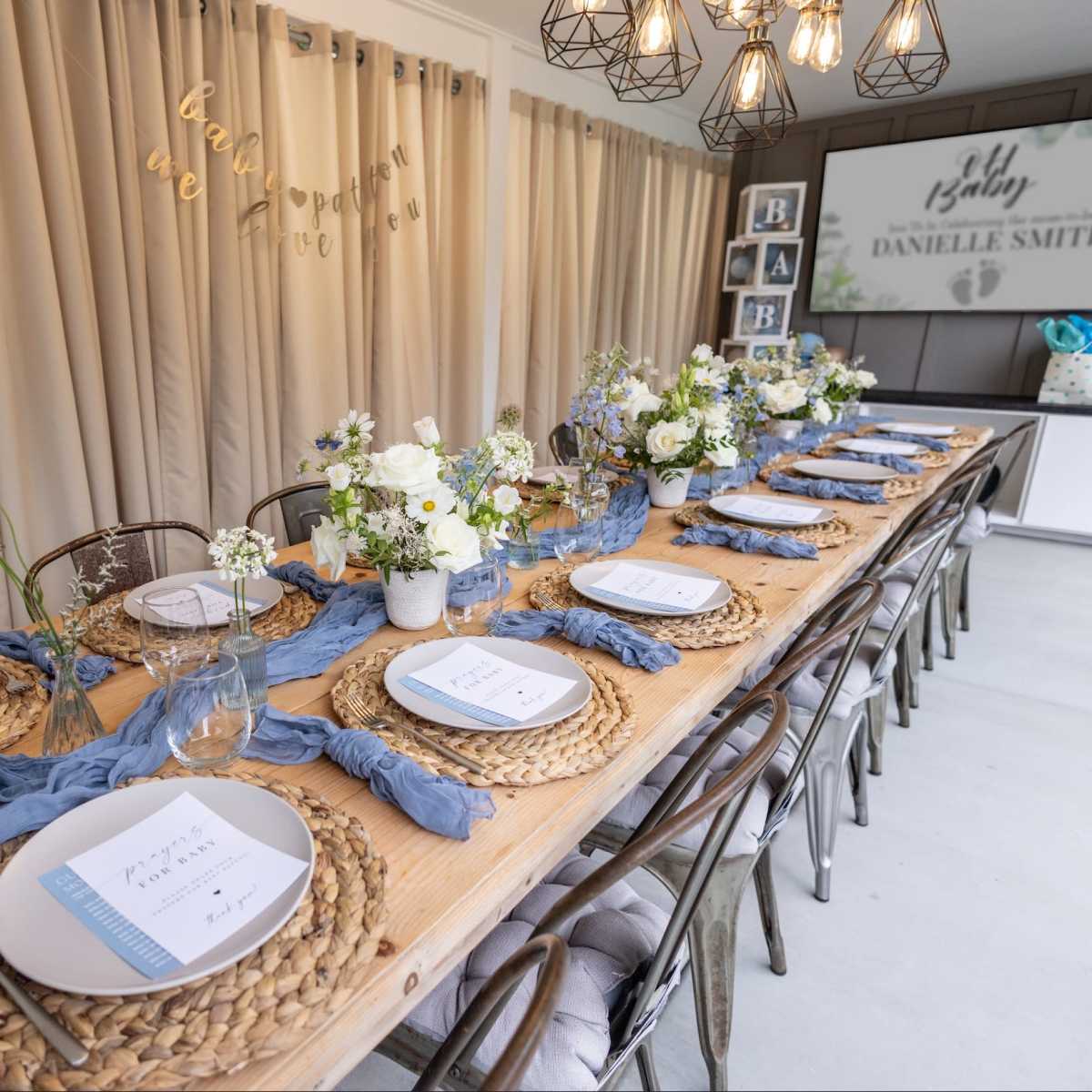 A long wooden table is set for a gathering, adorned with white flowers, blue runner fabric, and name cards.