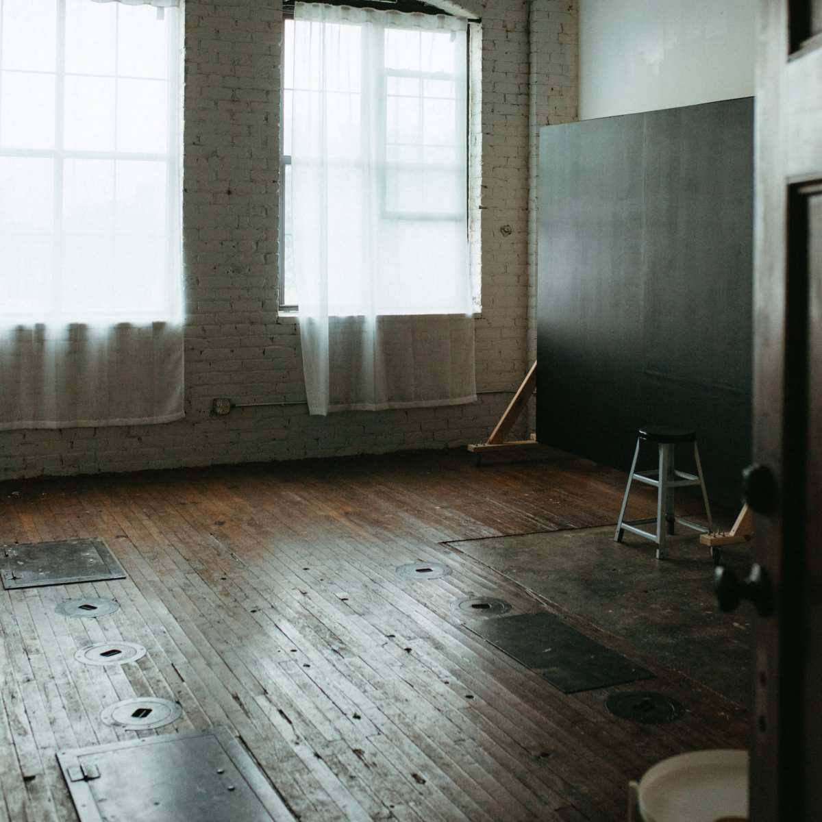 An empty room with wooden flooring, large windows draped with white curtains, and a tall stool positioned near a wall.