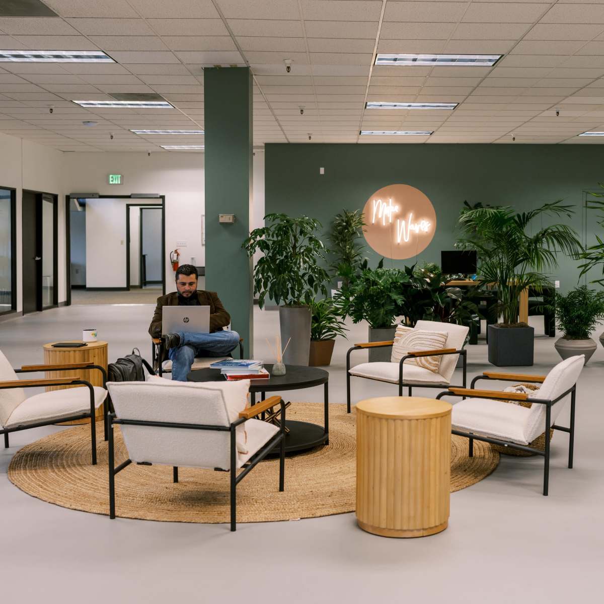 A man works on his laptop in a modern, well-lit office space surrounded by plants and comfortable seating.