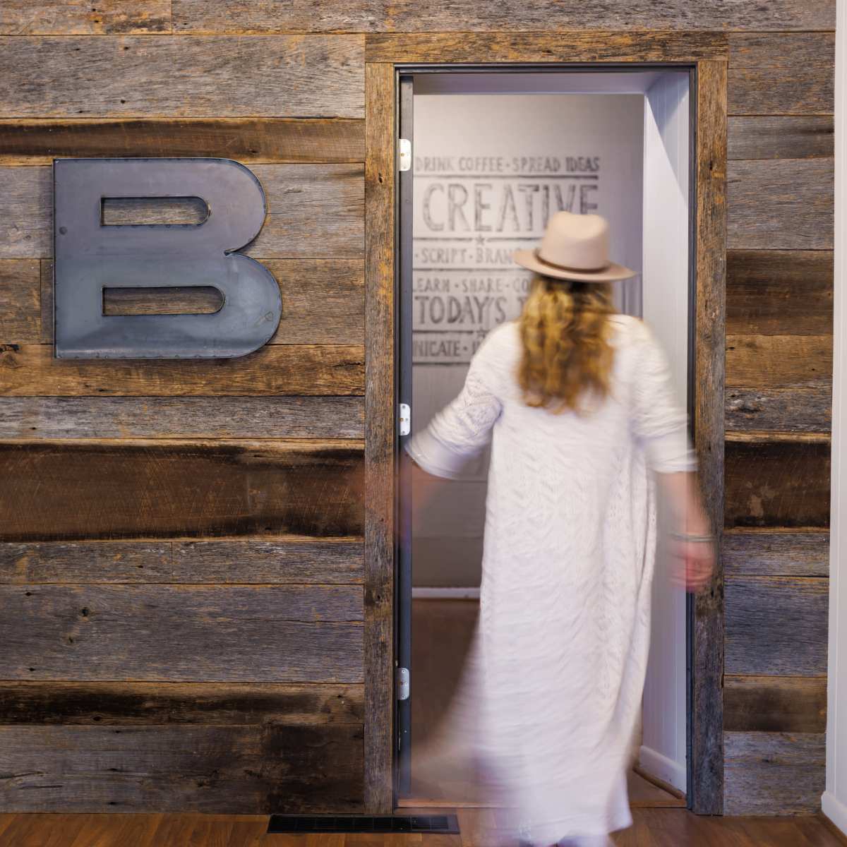 A person in a long white dress walks towards a partially open door with a metallic letter "B" on a rustic wooden wall.