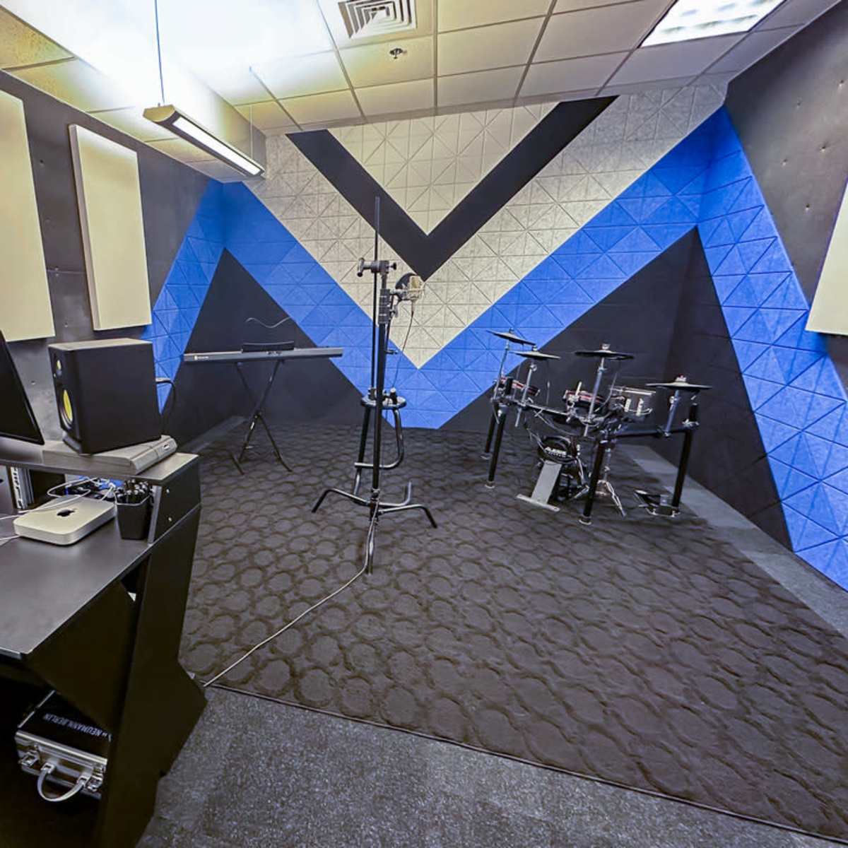 A music recording studio features a computer setup, a keyboard, and electronic drums against a patterned blue and black wall.