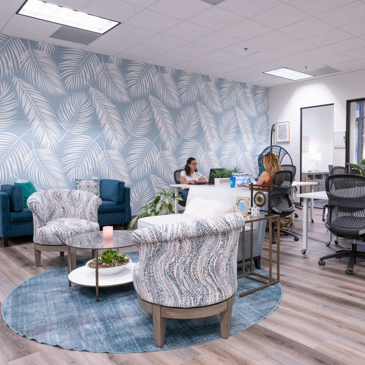 A modern office space featuring blue and white patterned wallpaper, a circular rug, and various seating arrangements including chairs and desks, with two women engaged in conversation.