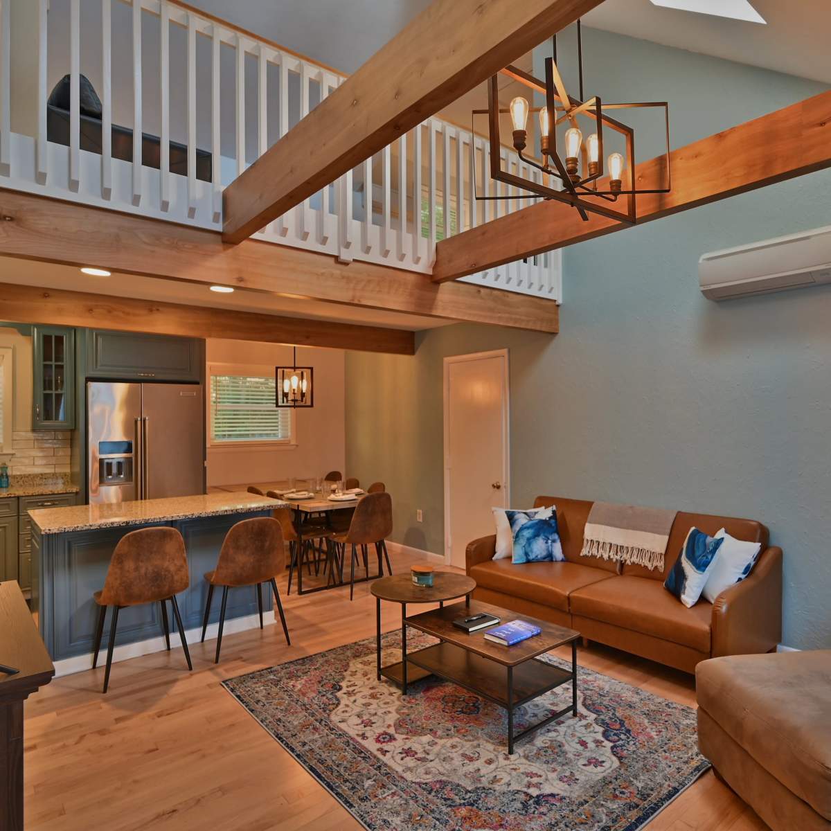 A modern living room with a brown sofa, a coffee table, and an open kitchen area featuring bar stools and a dining set beneath wooden beams.