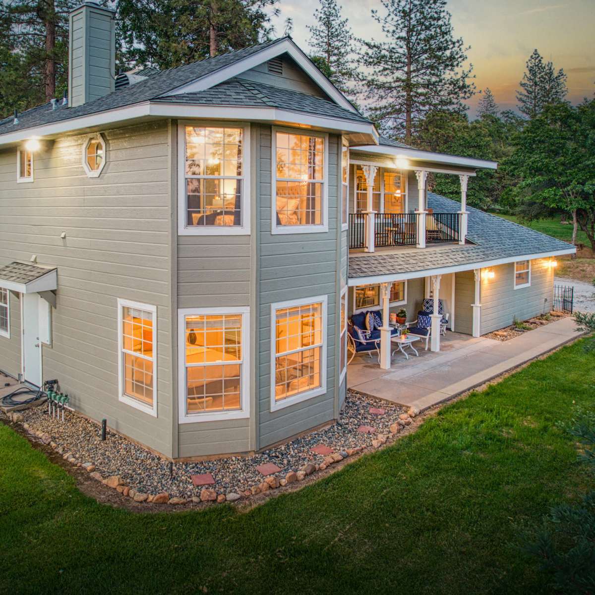 A two-story house with large windows, surrounded by greenery and a patio area, is illuminated in the evening light.