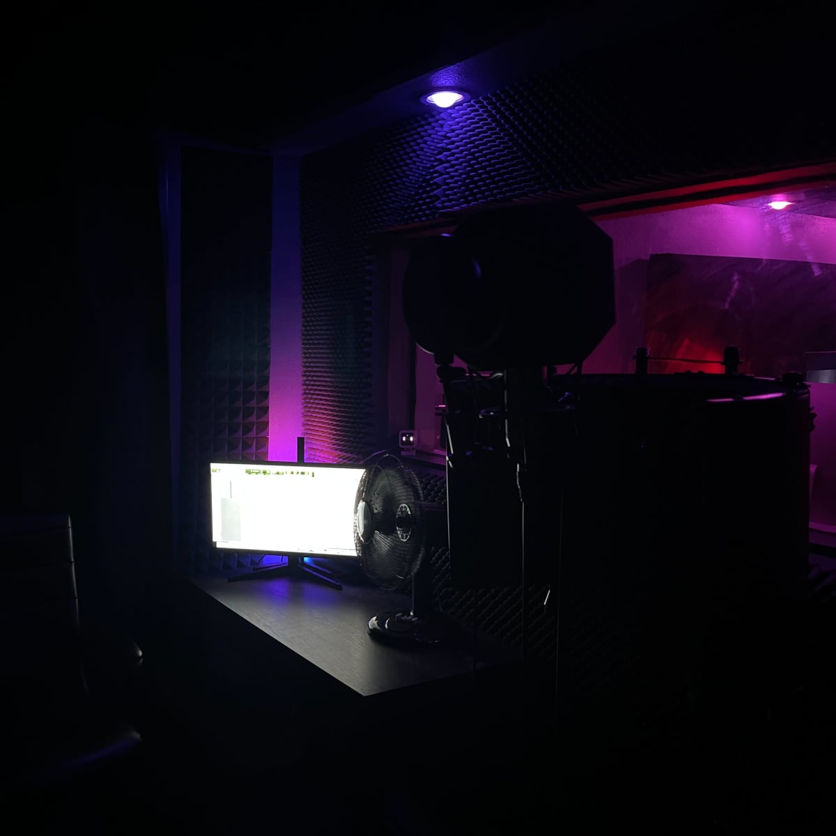 A dimly lit recording studio with a desk, a computer monitor, a fan, and a microphone setup against a backdrop of soundproofing panels.