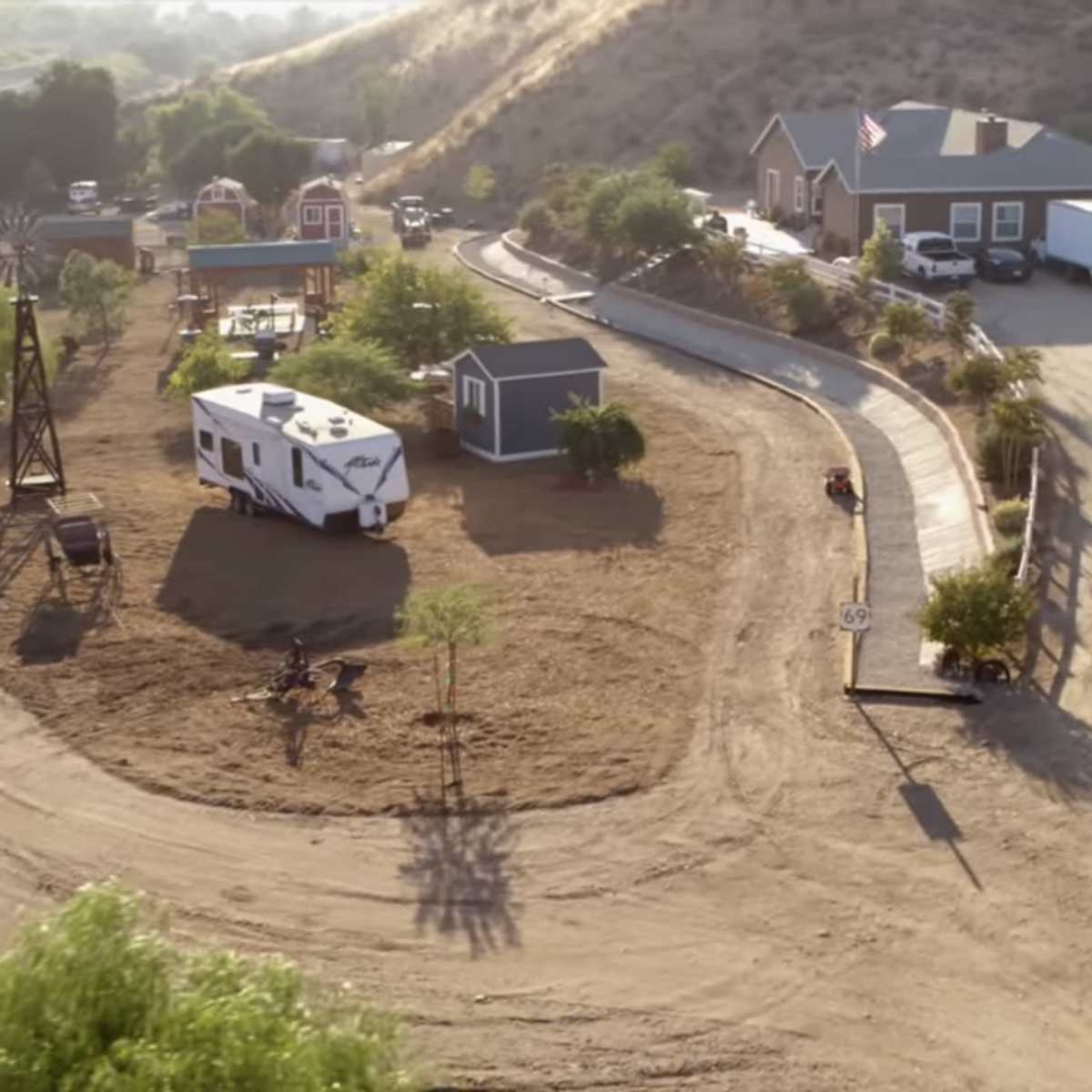 A rural property with a winding dirt driveway, a recreational vehicle parked among several trees, and a small house with additional structures in a hilly landscape.