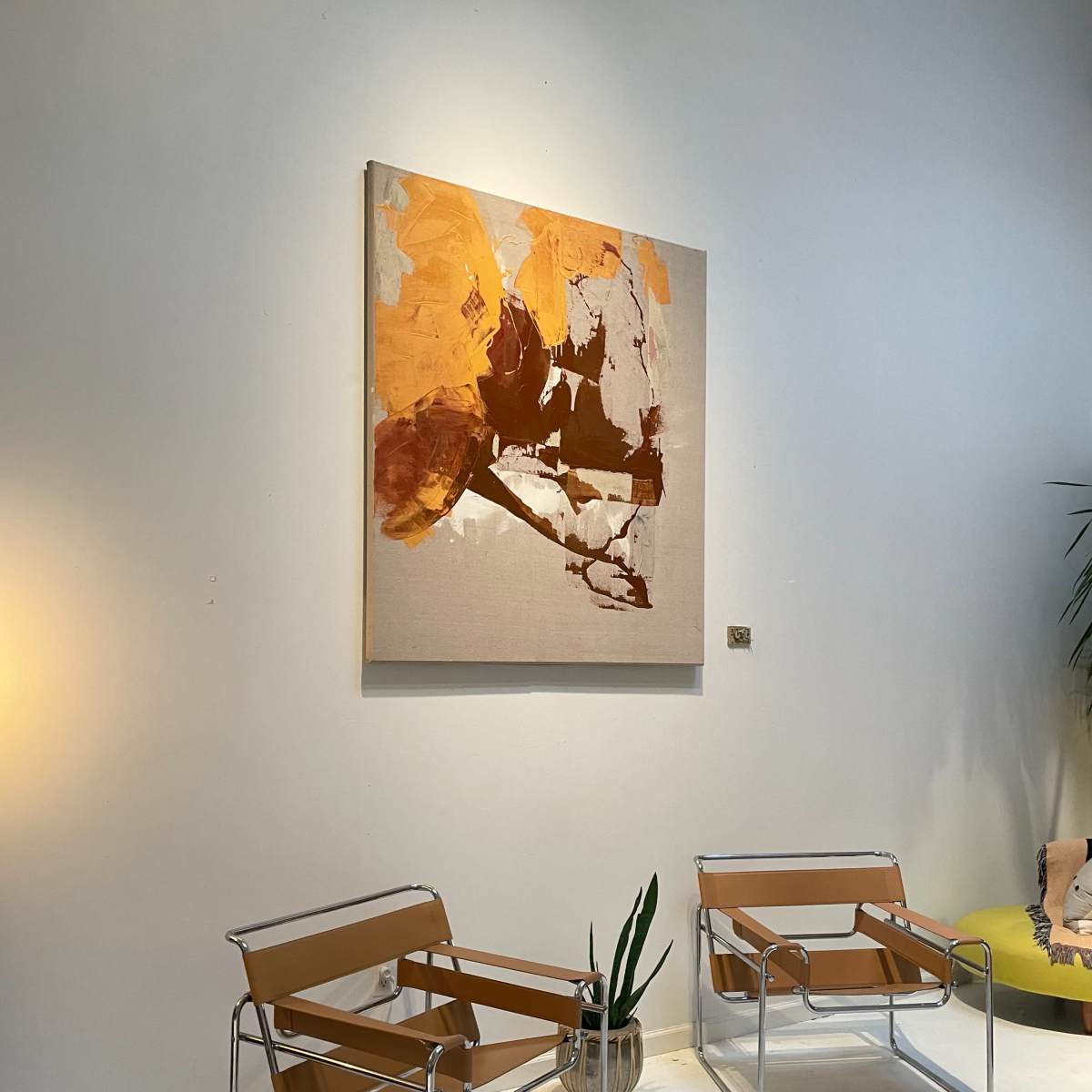 Two metal-framed chairs with tan fabric sit opposite a large abstract painting in warm tones on a pale wall, accompanied by a potted plant nearby.