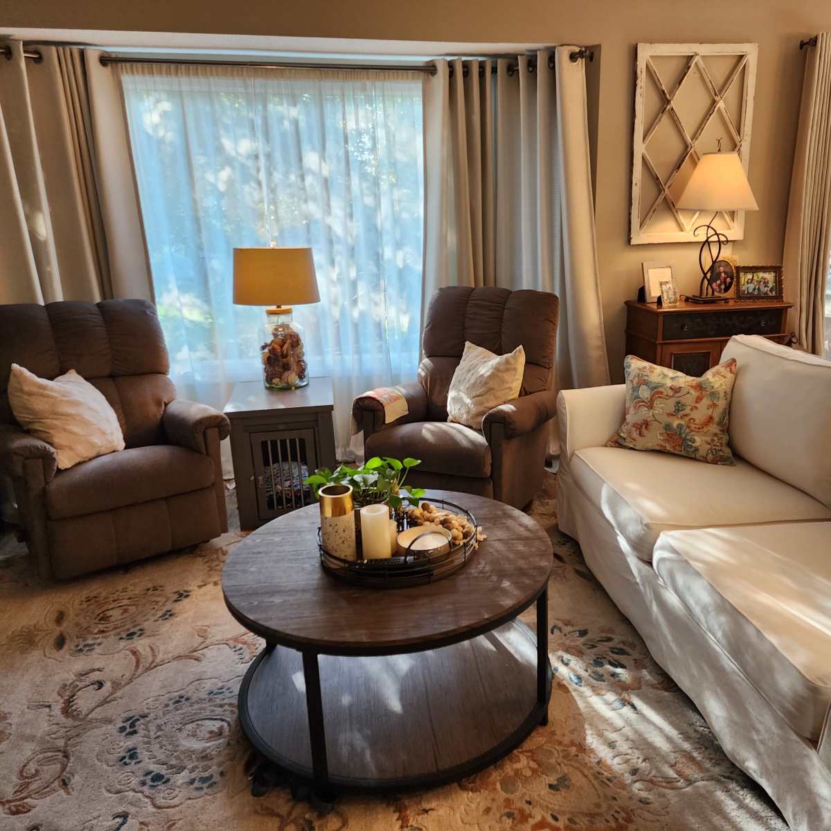 A cozy living room with two recliners, a light-colored sofa, a round coffee table adorned with decorative items, and large windows letting in natural light.