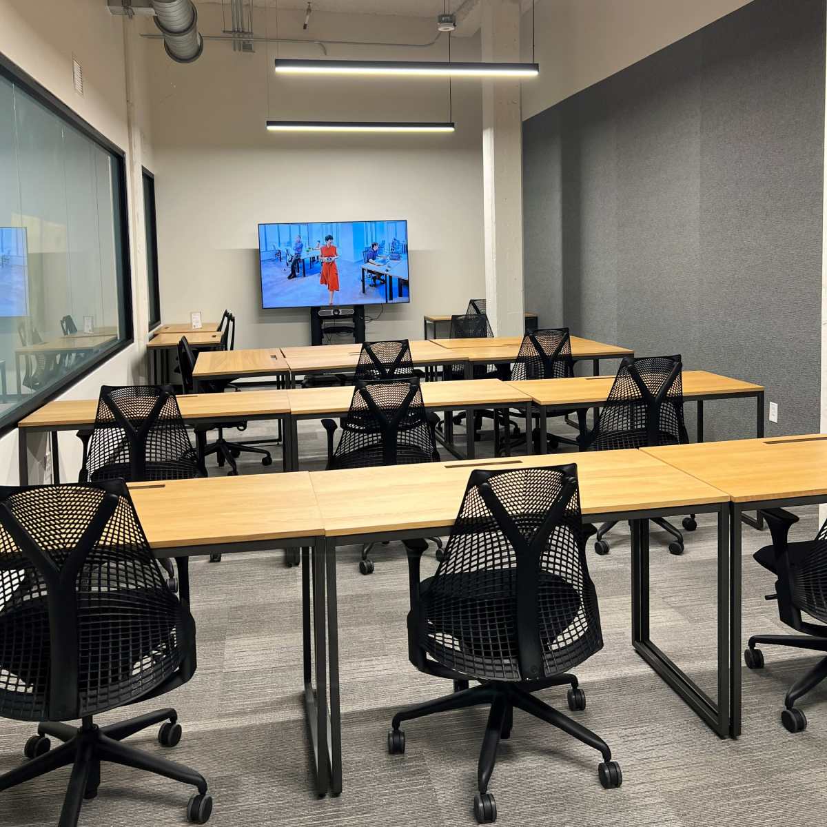 A modern conference room features multiple tables and chairs arranged with a large screen displaying a presentation on one wall.
