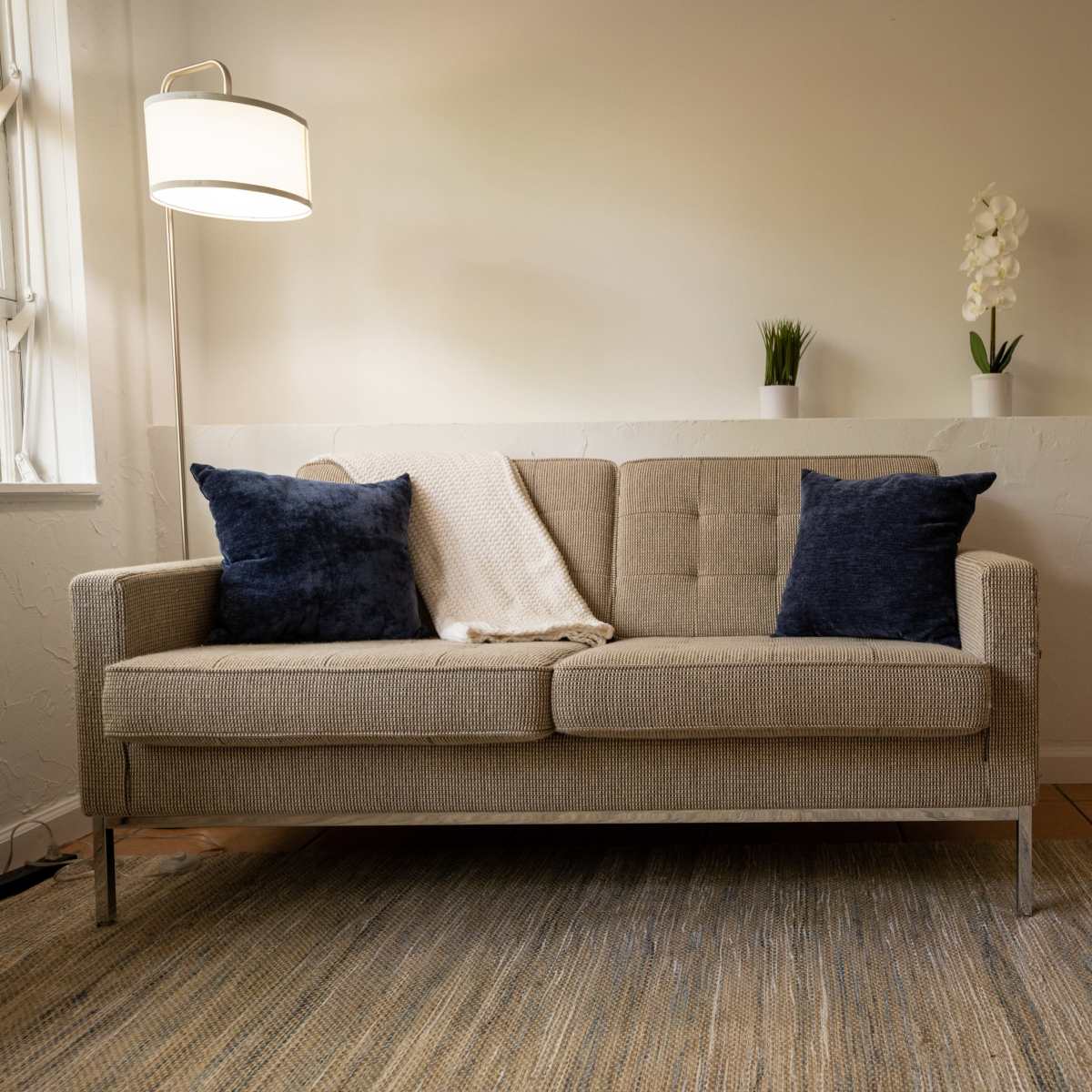 A light-colored sofa with two dark pillows and a throw blanket sits in a minimalist room with a lamp, potted plants, and a piece of artwork on the wall.