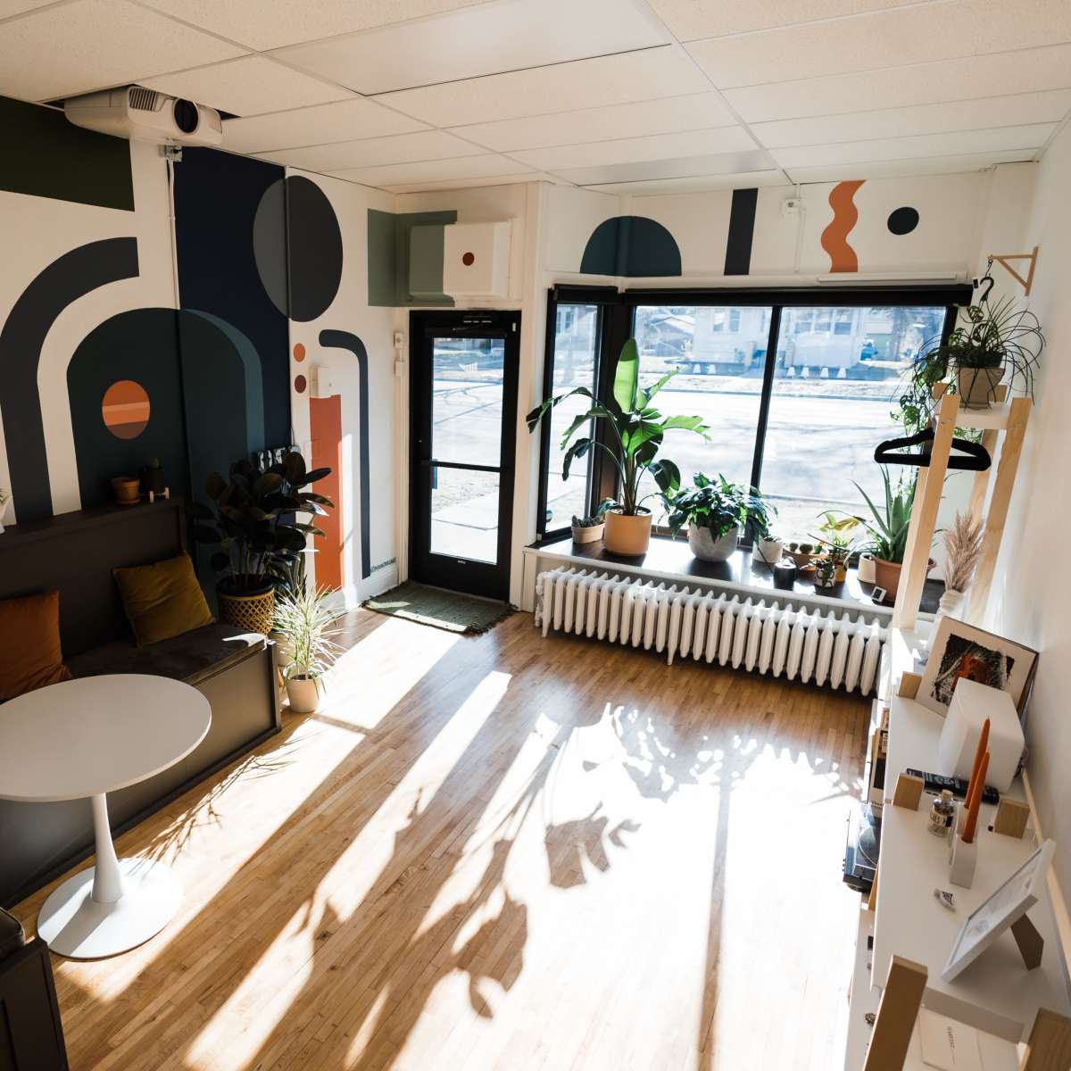 A modern, brightly lit room with a sectional sofa, a round table, and large windows displaying potted plants and geometric wall art.