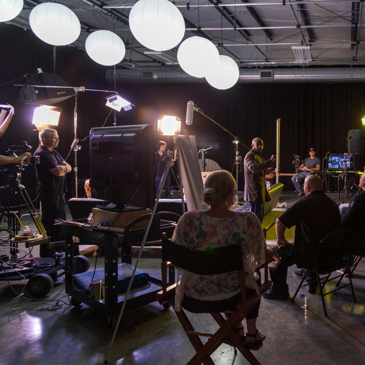 A group of people sits in chairs facing a presenter in a well-lit studio equipped with various cameras and lighting equipment.