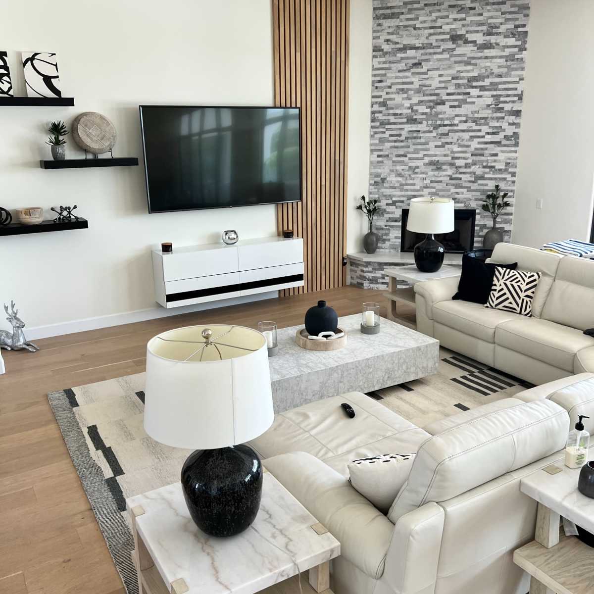 A modern living room with a large flat-screen TV mounted on the wall, a stone feature wall, light-colored furniture, and decorative elements on the shelves.