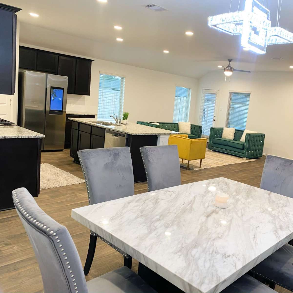 A modern dining area with a marble table surrounded by upholstered chairs, leading into an open-concept living space featuring a kitchen and a sitting area with colorful couches.