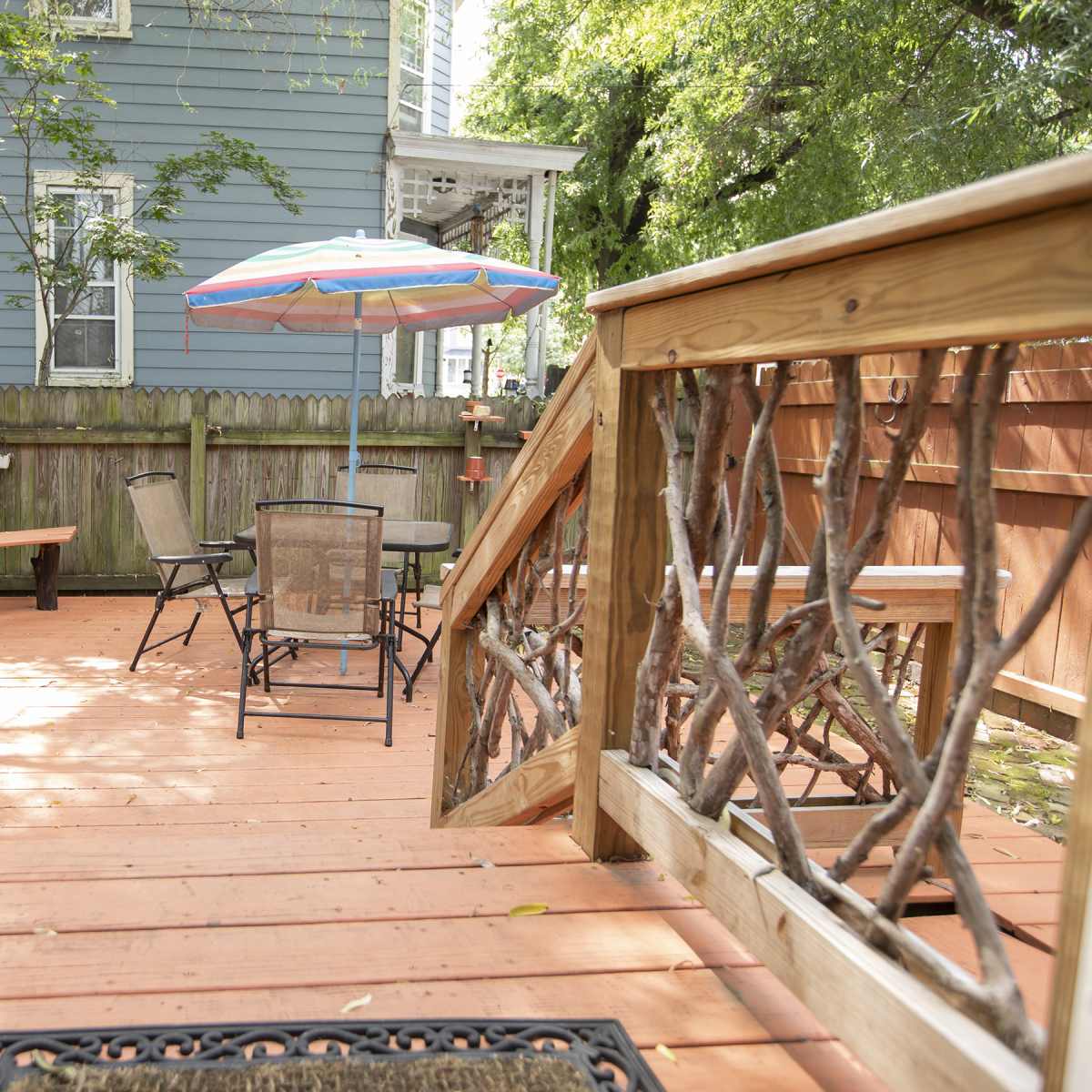 A wooden deck with a hot tub, a chair and table set, and a colorful umbrella, surrounded by greenery and a blue house in the background.