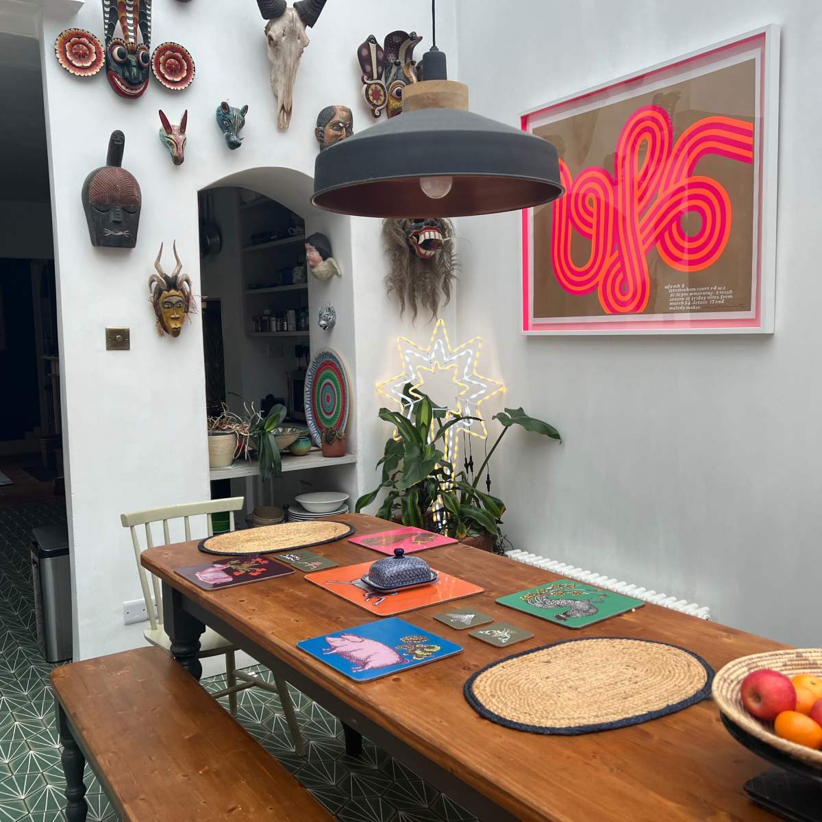 A wooden dining table with colorful placemats is set under a pendant light in a room adorned with various masks and artwork on the walls.