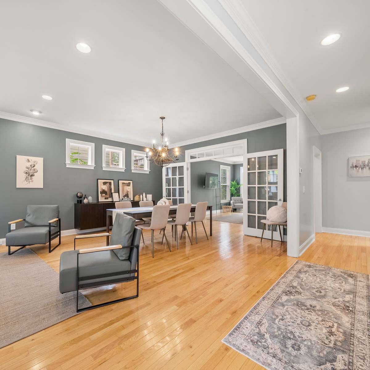 The room features a light wood floor, a dining area with a table and chairs, a sitting area with armchairs, and large windows allowing natural light.