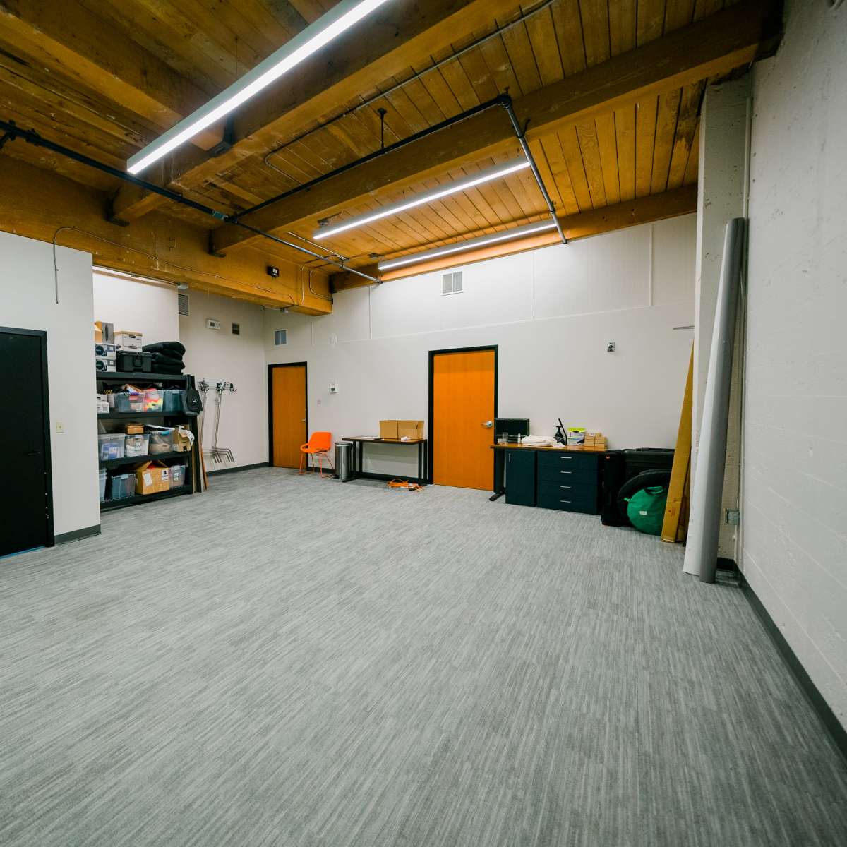 A spacious, empty room with a wooden ceiling, gray carpet, and minimal furniture including a desk and orange chair.