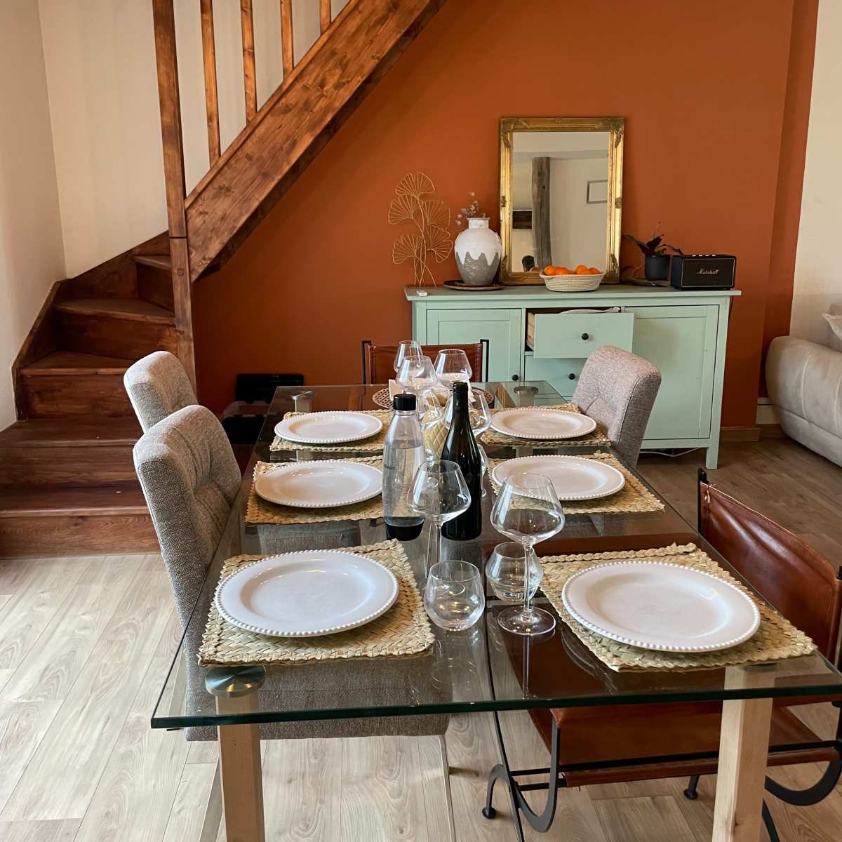 A dining table is set with plates and glassware, positioned near a staircase and a sideboard against an orange wall.