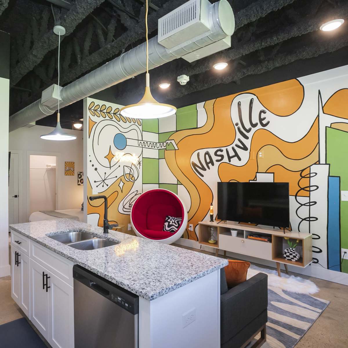 A modern kitchen and living area feature a vibrant mural reading "Nashville" on one wall, with a red accent chair and contemporary furnishings.