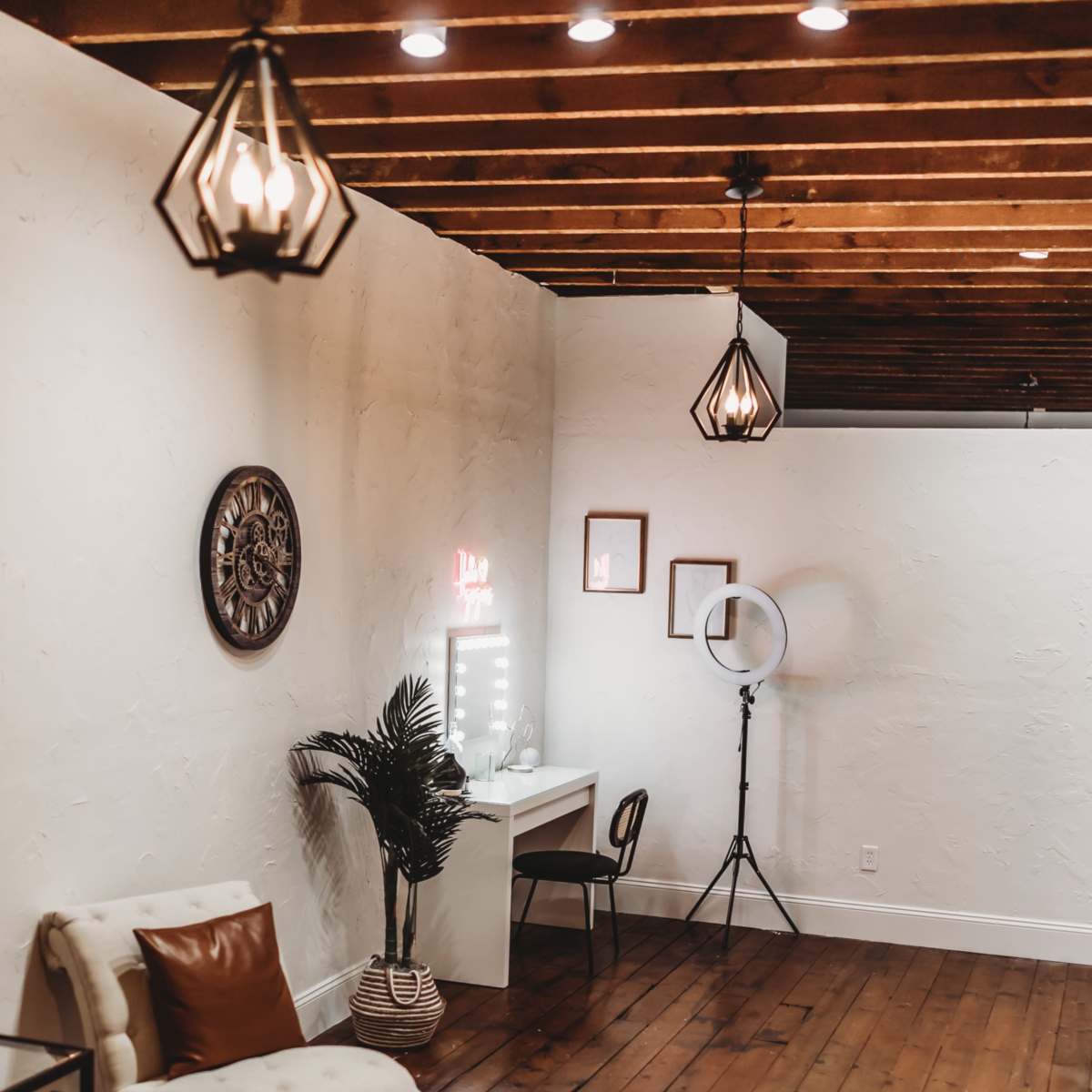 A well-lit room with wooden flooring, featuring a desk with a mirror, a comfortable chair, decorative lighting, and a potted plant.