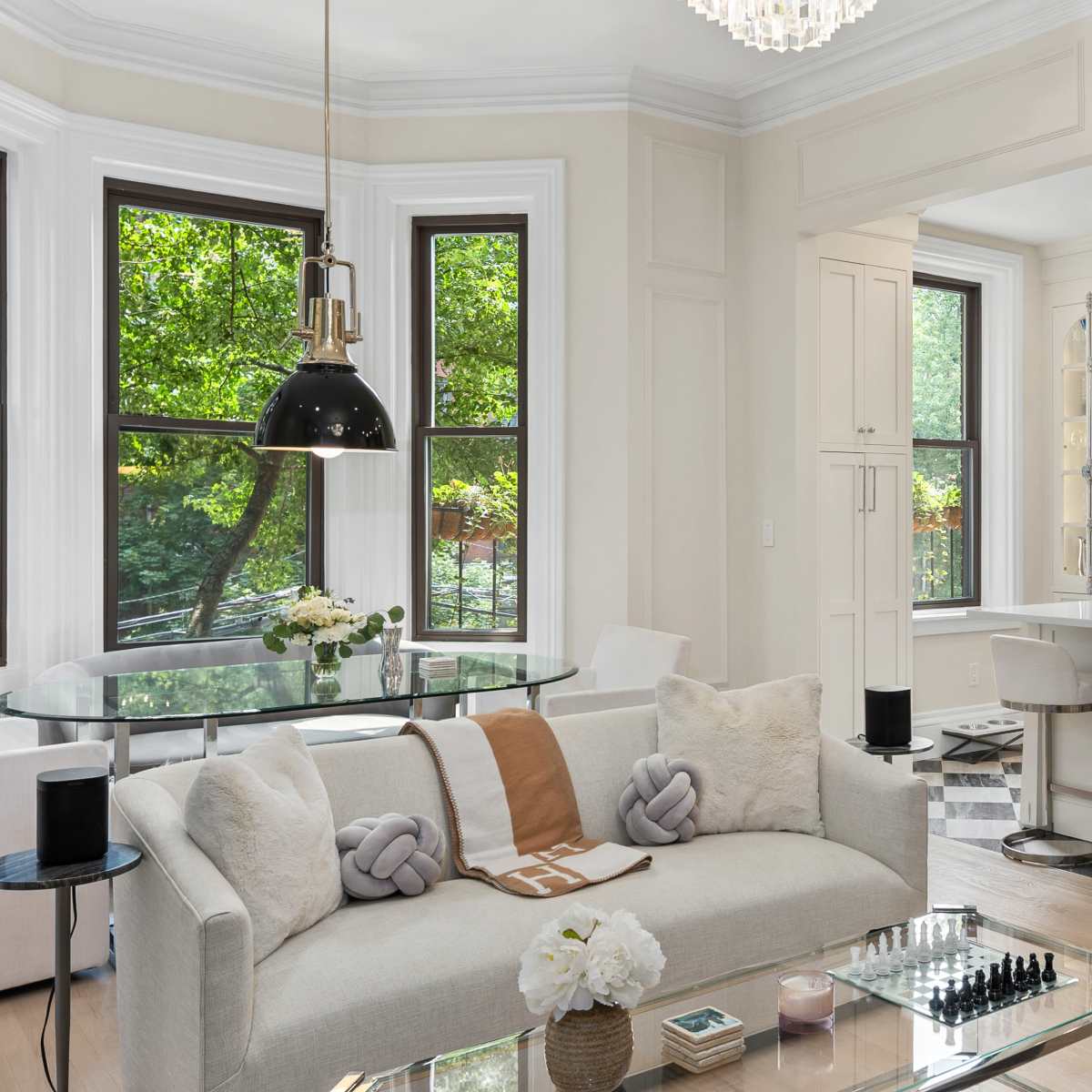 A modern living space features a light-colored sofa, a glass dining table, and a kitchen with wooden shelves displaying decorative items.