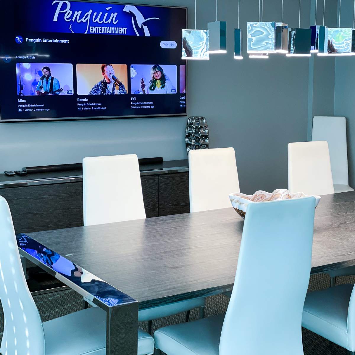 A modern conference room features a long table surrounded by light blue chairs, with a TV displaying "Penguin Entertainment" on the wall.