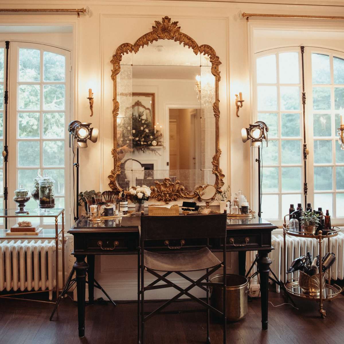 A vintage vanity table with a large ornate mirror is positioned between two windows, surrounded by makeup tools and lighting equipment.