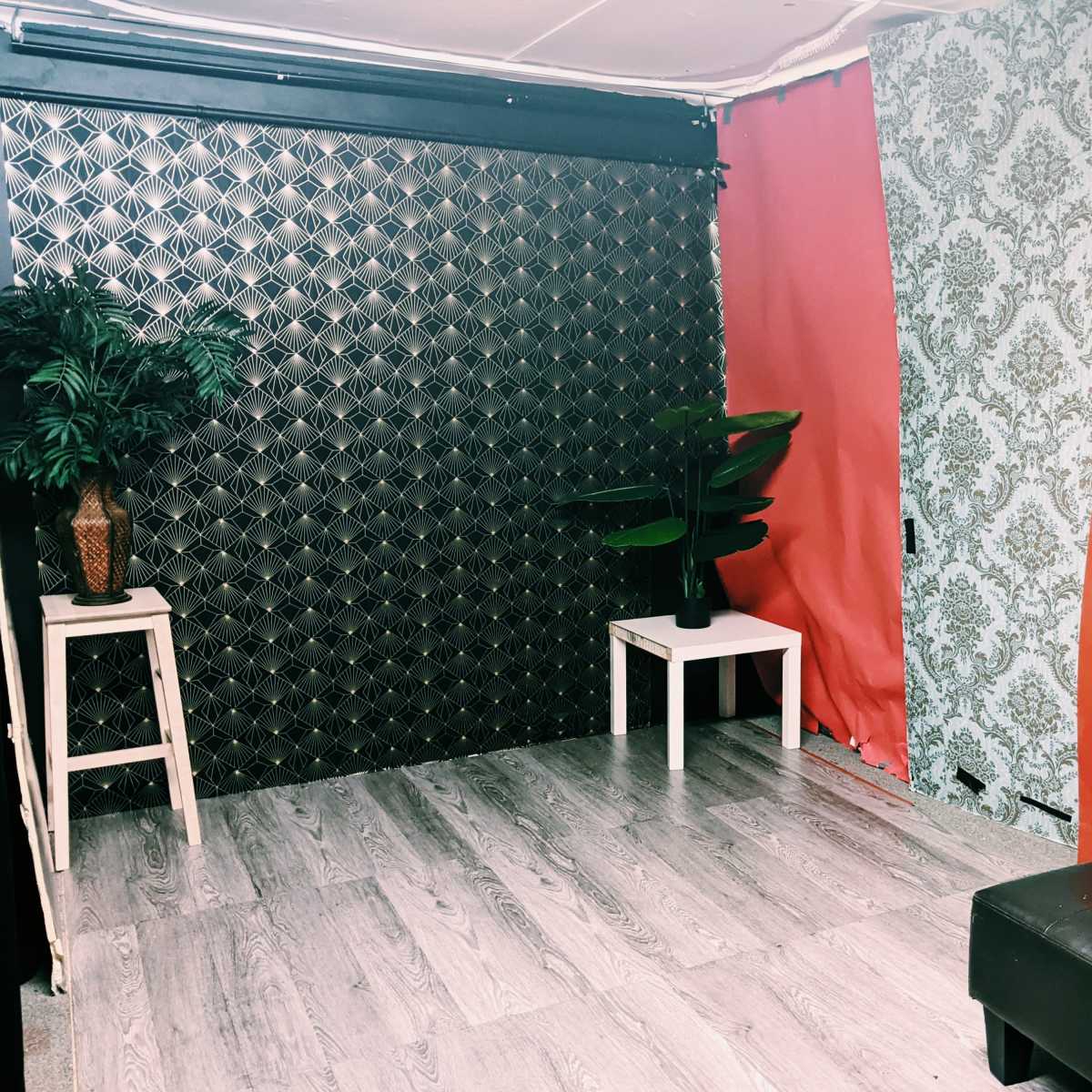 A small, decorated room with a black textured wall, a red fabric wall, a floral blue wall, a gray couch, and a small white table with a plant.