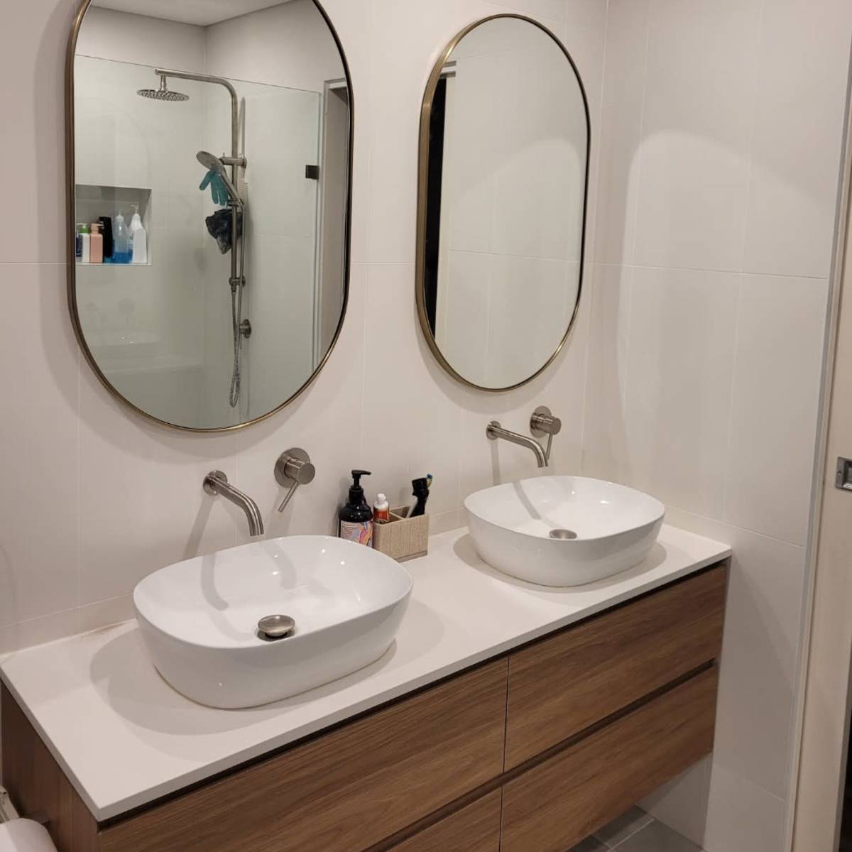 A modern bathroom with two oval mirrors above separate sinks on a wooden vanity, and a shower area visible in the background.