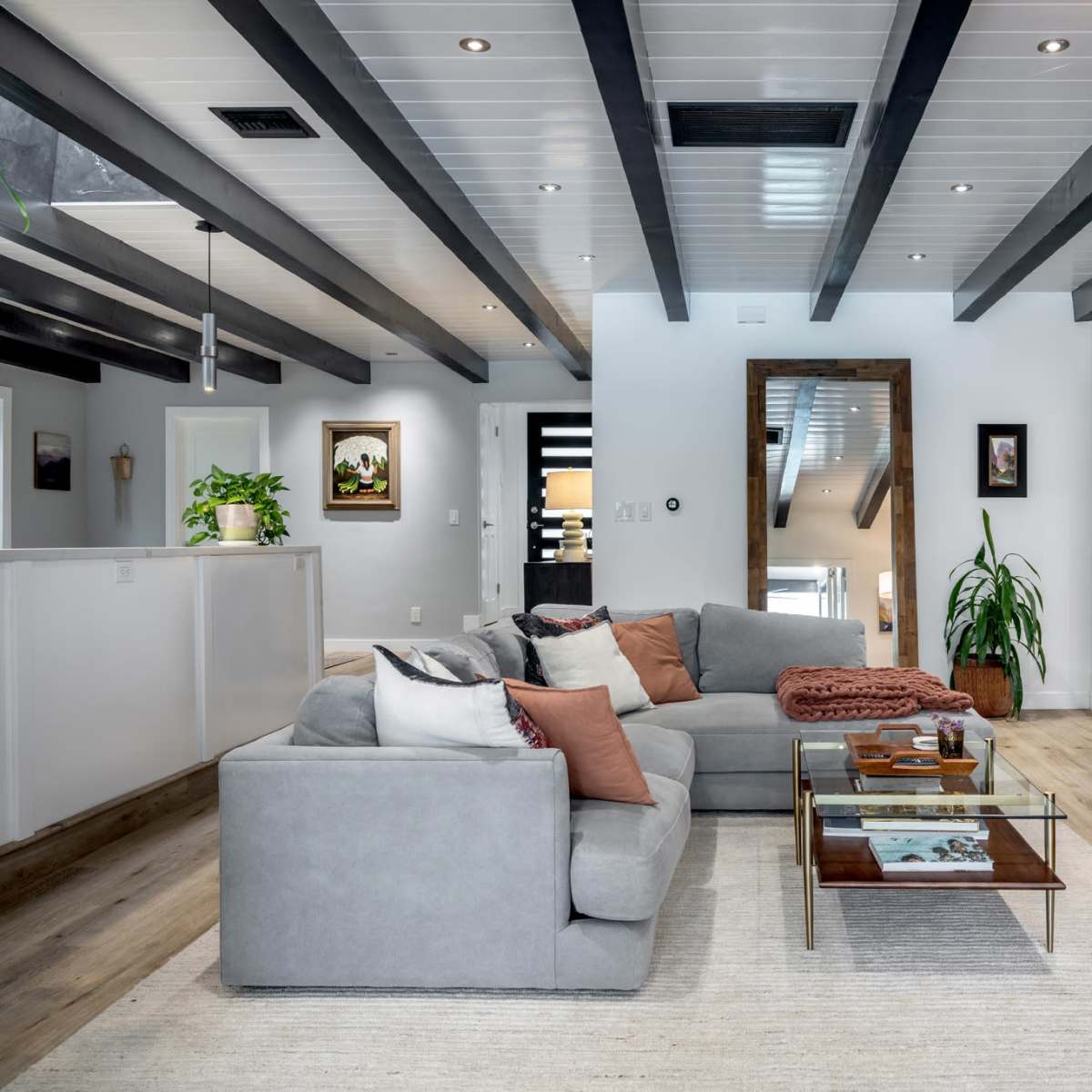 The living room features a gray sectional sofa, a glass coffee table, wooden shelving with decor, and large windows with a view of greenery.