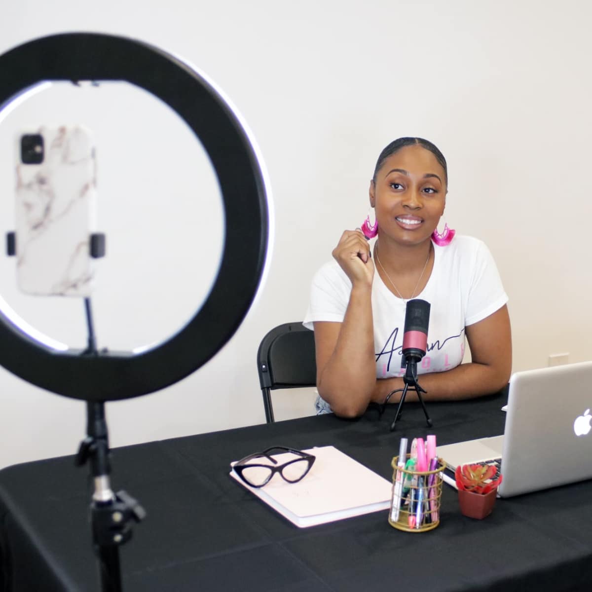 A woman with a ponytail sits at a desk with a laptop, a ring light, and various office supplies.