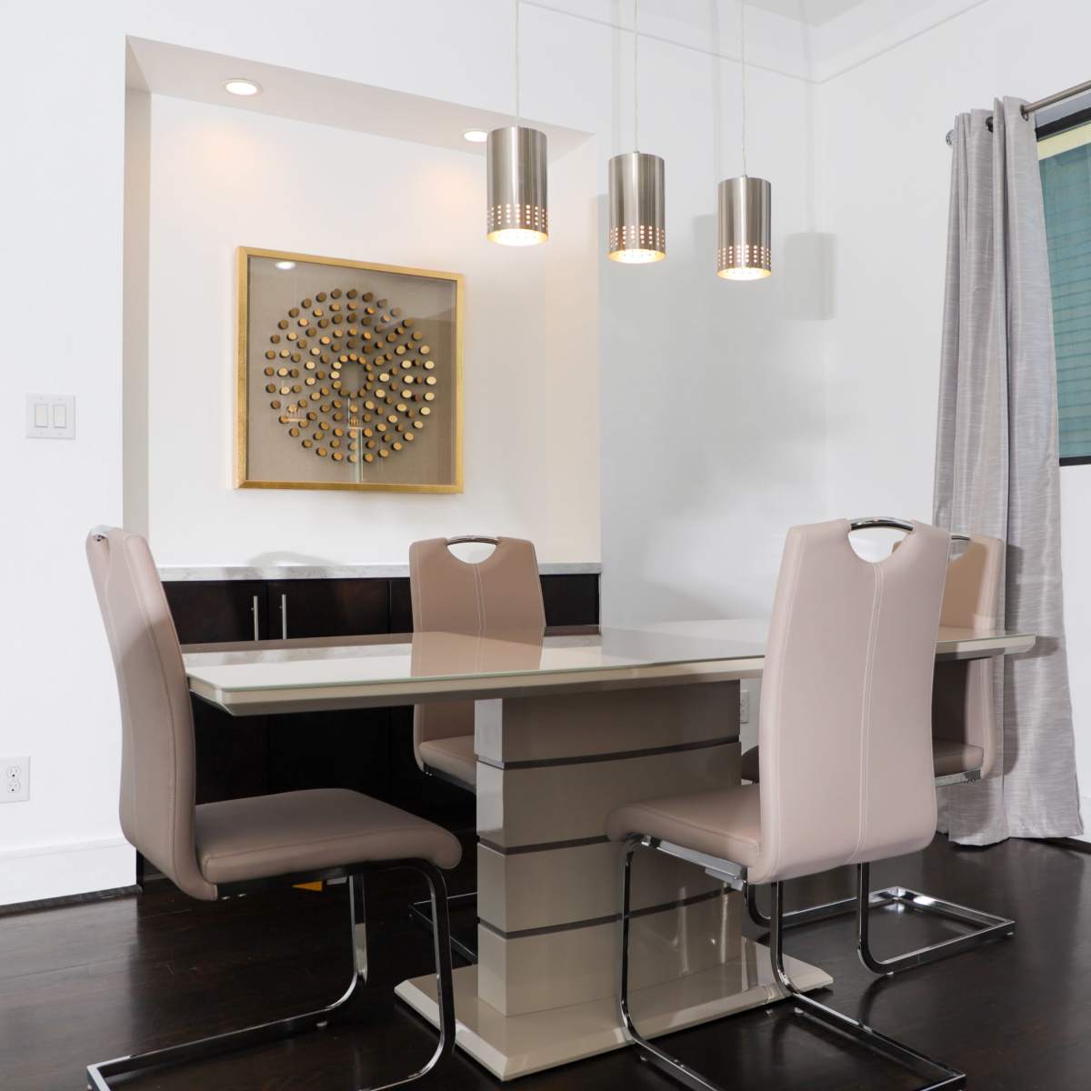 A modern dining area featuring a rectangular table with four upholstered chairs, situated under three pendant lights, alongside a wall with a decorative circular wall art.