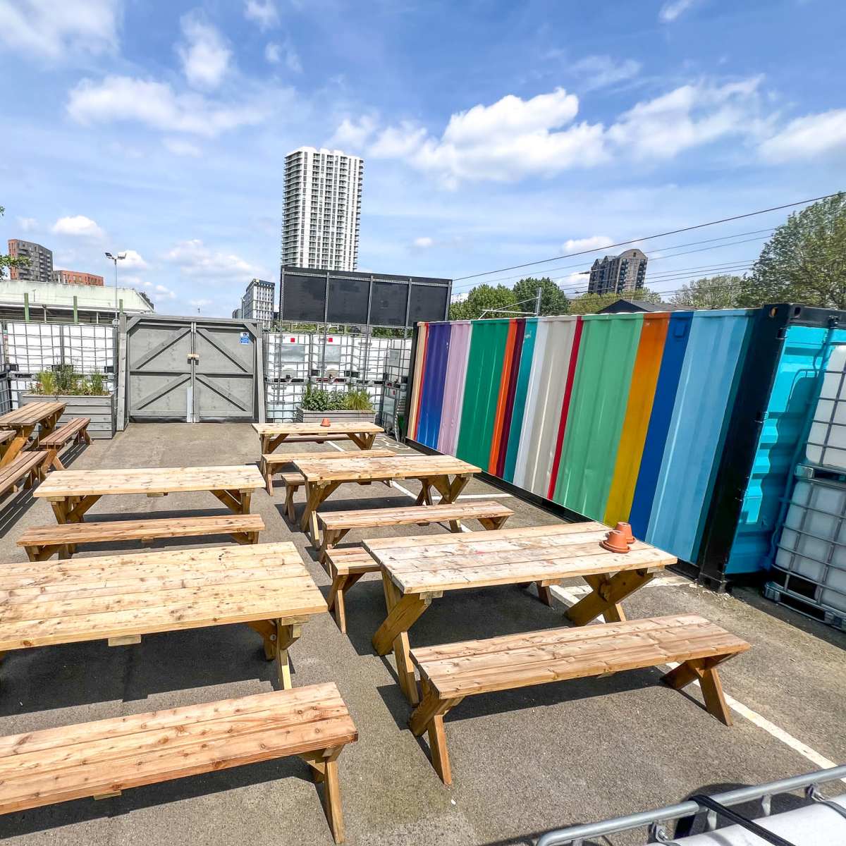 An outdoor seating area with wooden picnic tables arranged in rows, surrounded by storage containers of various colors and a view of tall buildings in the background.