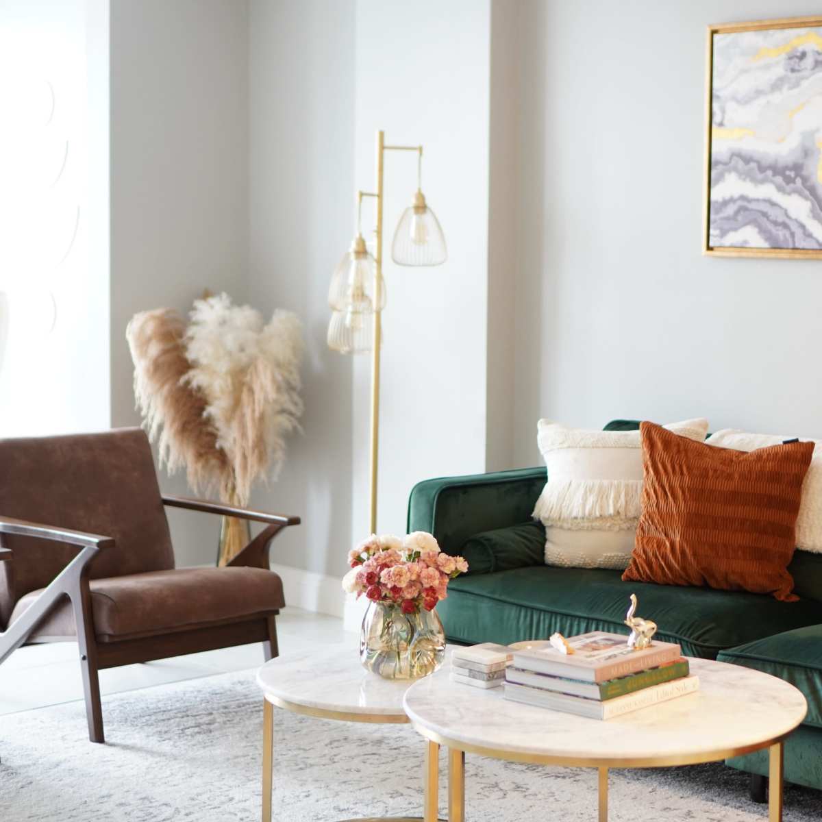 A modern living room features a green velvet sofa with decorative pillows, a marble coffee table, and a stylish artwork above, complemented by a floor lamp and floral arrangements.