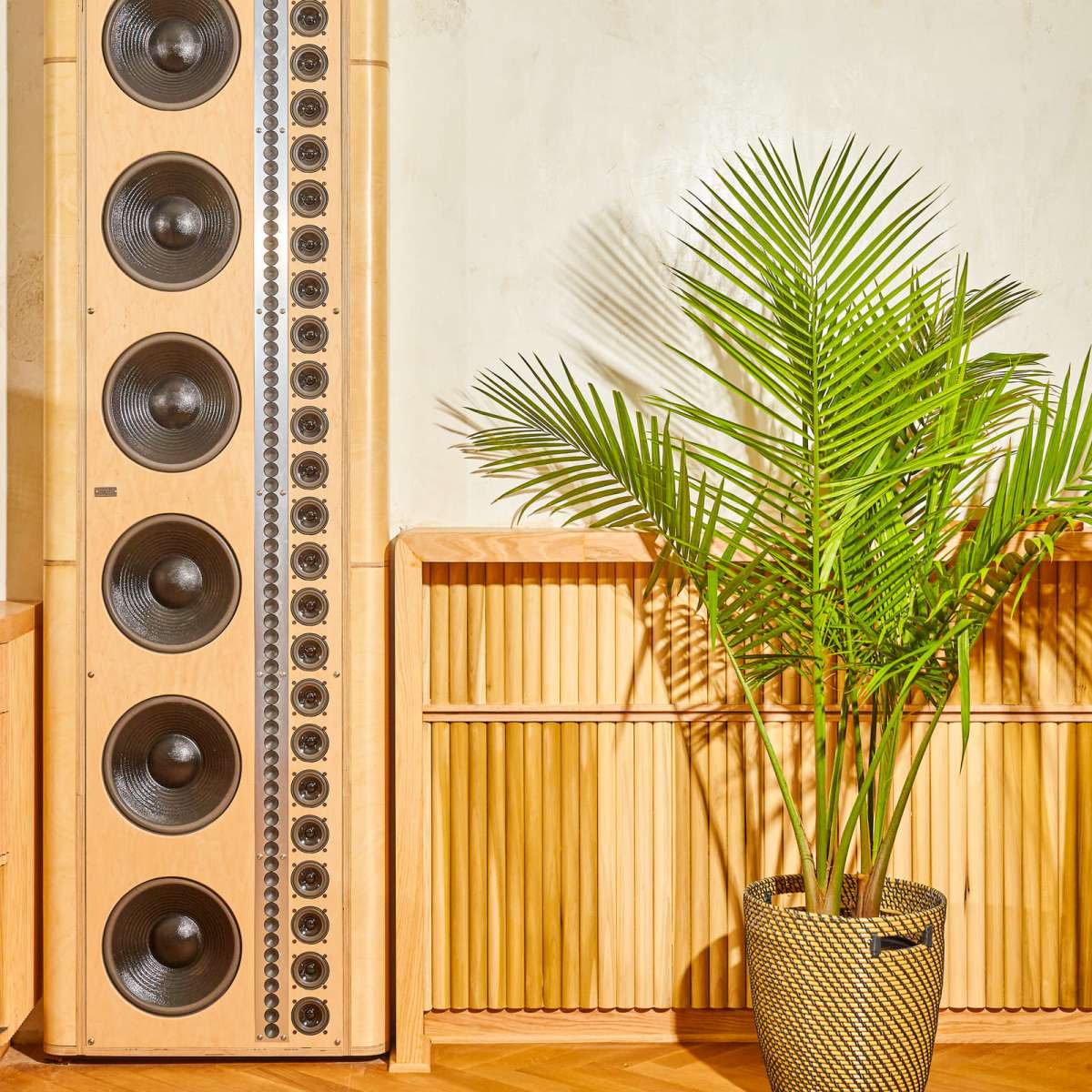 A tall speaker with multiple woofers stands next to a potted palm plant in a room with a textured wall.