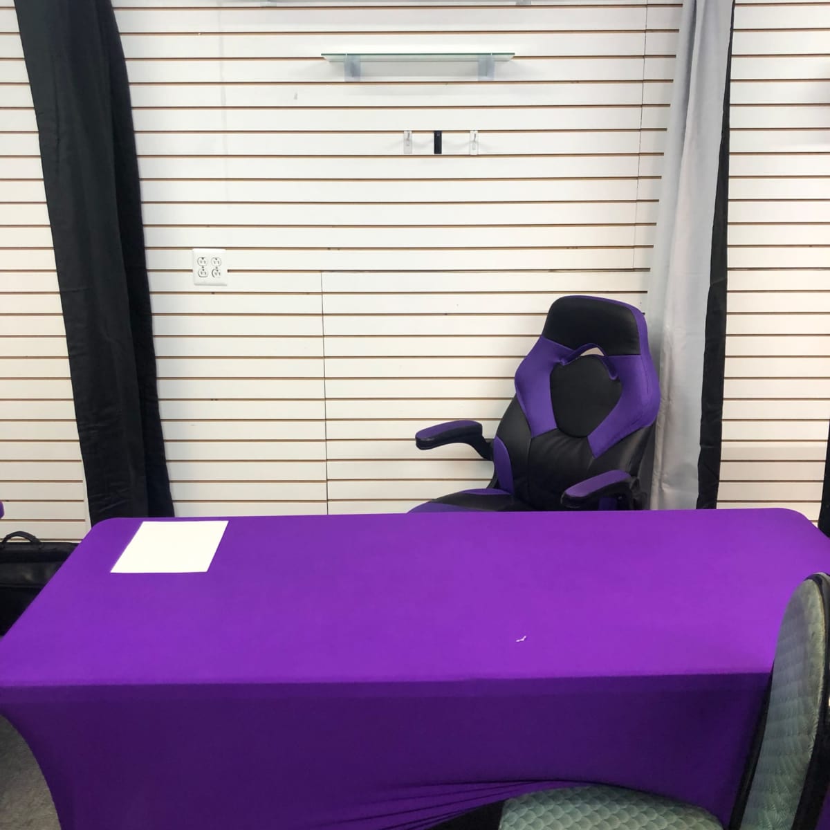A purple table is situated in front of a black and white backdrop with a shelf above it and an ergonomic chair beside the table.