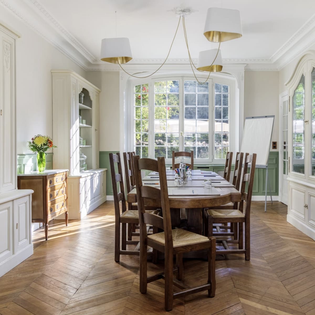 A wooden dining table surrounded by chairs is situated in a well-lit room with large windows, decorative cabinetry, and a herringbone-patterned floor.