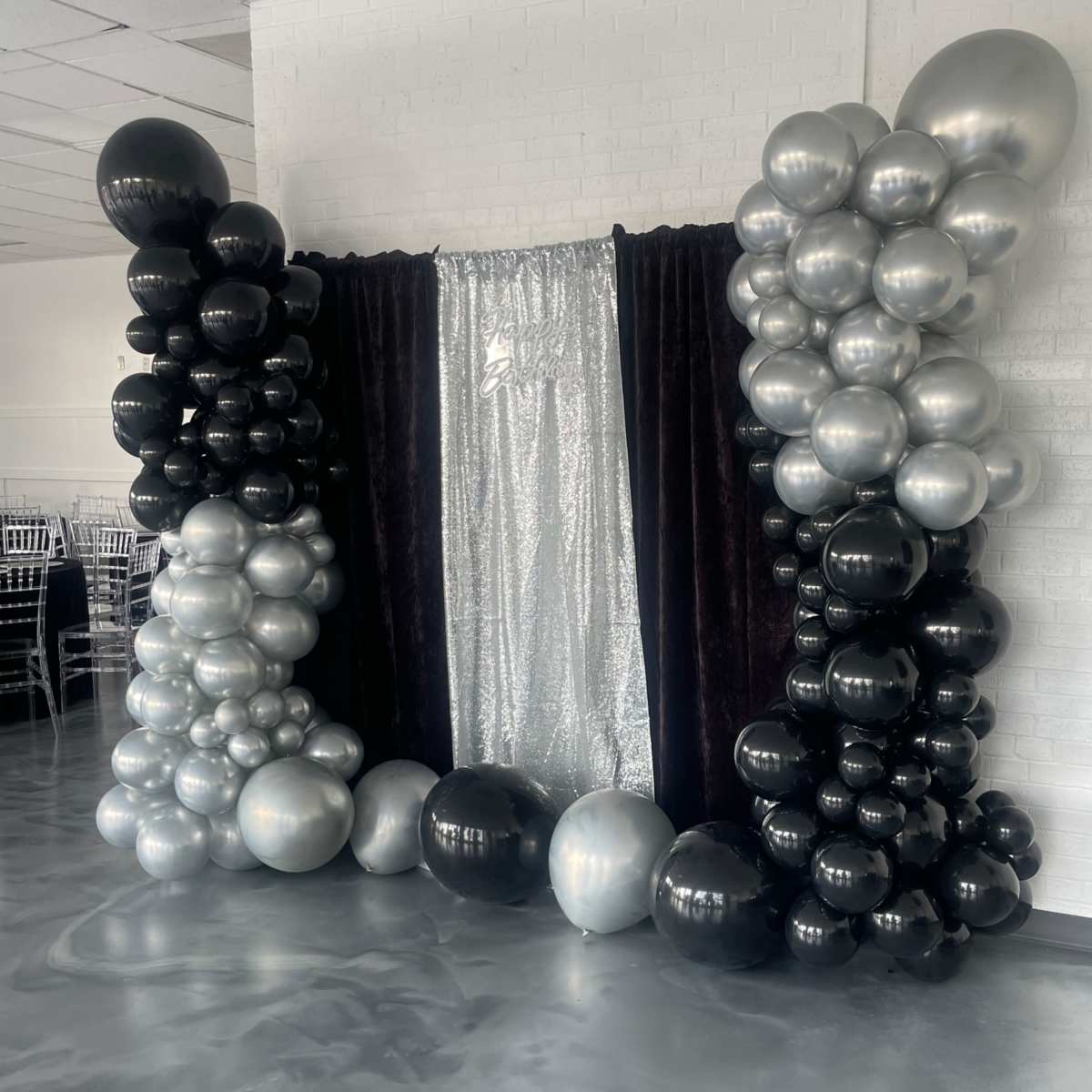 A decorative backdrop for an event, featuring a curtain in the center surrounded by clusters of black and silver balloons.