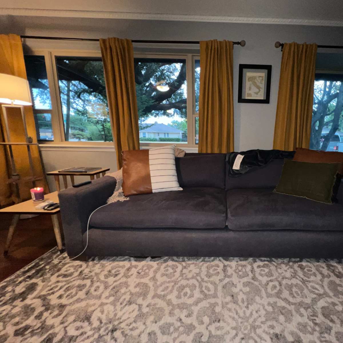 A dark sofa is positioned in a living room with yellow curtains and a patterned rug, near large windows that overlook a yard.