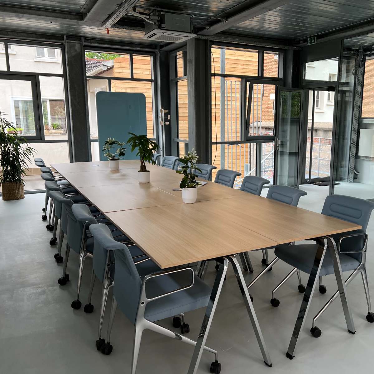 A modern conference room with a long wooden table surrounded by blue office chairs and large windows allowing natural light.