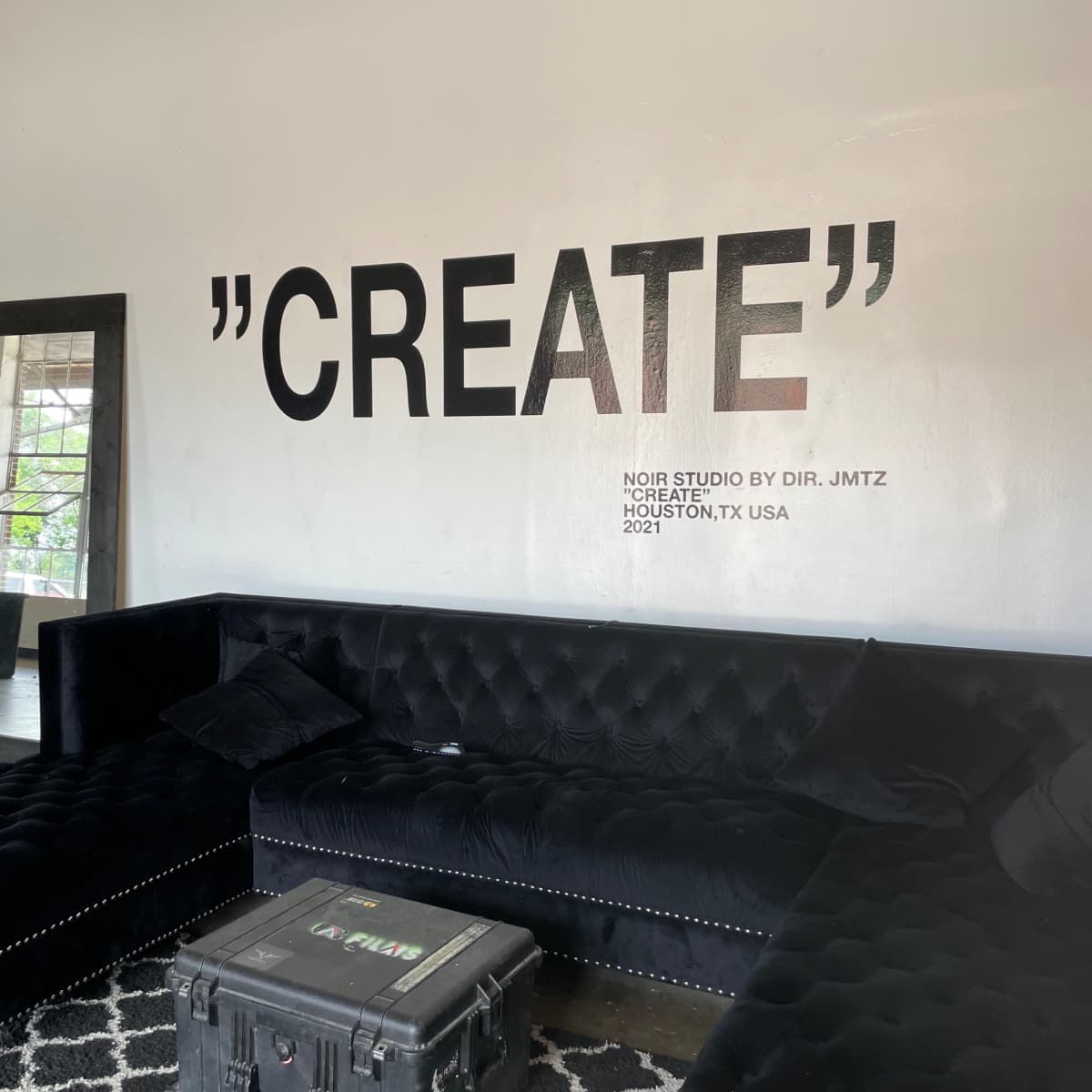 A modern lounge area featuring a black couch, a full-length mirror, and a wall with the word "CREATE" prominently displayed in large letters.